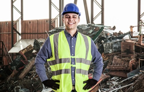 A male waste management professional is standing inside a large industrial warehouse, surrounded by a pile of discarded metal and wooden debris. He is wearing a blue safety helmet, black gloves, and a high-visibility yellow safety vest over a blue shirt. The debris includes pieces of rusted metal, broken wooden planks, and various scrap materials, indicating a rubbish clearance operation. The warehouse has a high ceiling with metal support beams and large windows allowing natural light to illuminate the scene. In the background, parts of a waste-truck or loading equipment are partially visible, suggesting an active rubbish removal site. The worker is smiling confidently, conveying professionalism and readiness to undertake clearance tasks. This image aligns with rubbish removal services offered by Earls Court House Clearance and demonstrates a typical scene of industrial waste management, likely in or near London, with the postcode area corresponding to Earls Court.