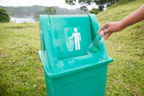 A person's hand is shown disposing of a clear plastic bottle into a bright turquoise recycling bin situated outdoors on a grassy area, with a background of trees, a lake, and hilly terrain under an overcast sky. The bin features a white icon of a person throwing trash into a bin, indicating it is designated for recyclable waste. The scene emphasizes waste disposal and recycling in an open environment, relevant to rubbish removal services in the local area near Earls Court, with the bin's vibrant colour contrasting against the green grass and natural surroundings. The gravel or pavement edge is slightly visible at the base of the bin, and the scene is well-lit with natural, diffuse daylight.