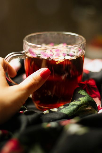 A close-up of a person's hand with neatly manicured nails painted red, holding a clear glass mug filled with a dark red herbal tea. The tea is topped with small, pink flower petals floating on its sur